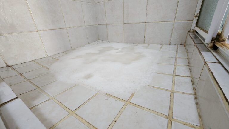 Water backing up into a tiled shower floor due to a blocked drain, causing wastewater to pool instead of draining away properly.