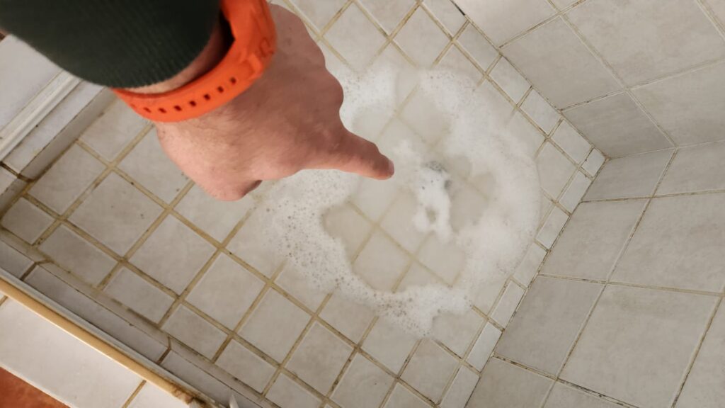 Hand pointing at standing water backing up around a shower drain, indicating a blocked shower drain and slow drainage problem.
