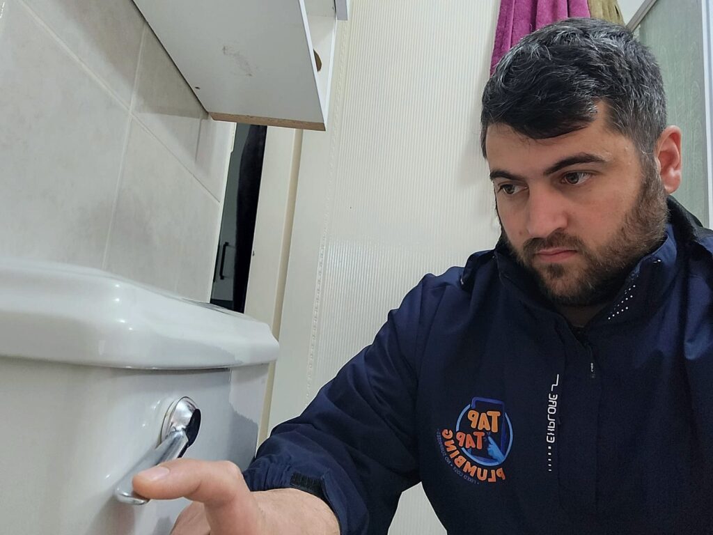 Tap Tap Plumbing technician inspecting a loose toilet handle in a homeowner's bathroom to diagnose the cause of continuous water flow and prevent water waste.